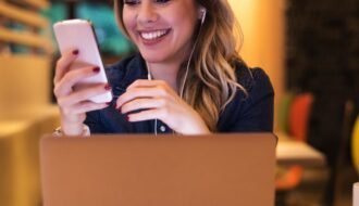 Mulher em frente ao laptop, com fones de ouvido, olhando o celular e sorrindo. Simboliza o sucesso em atrair clientes com o marketing no Instagram.