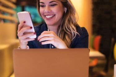 Mulher em frente ao laptop, com fones de ouvido, olhando o celular e sorrindo. Simboliza o sucesso em atrair clientes com o marketing no Instagram.